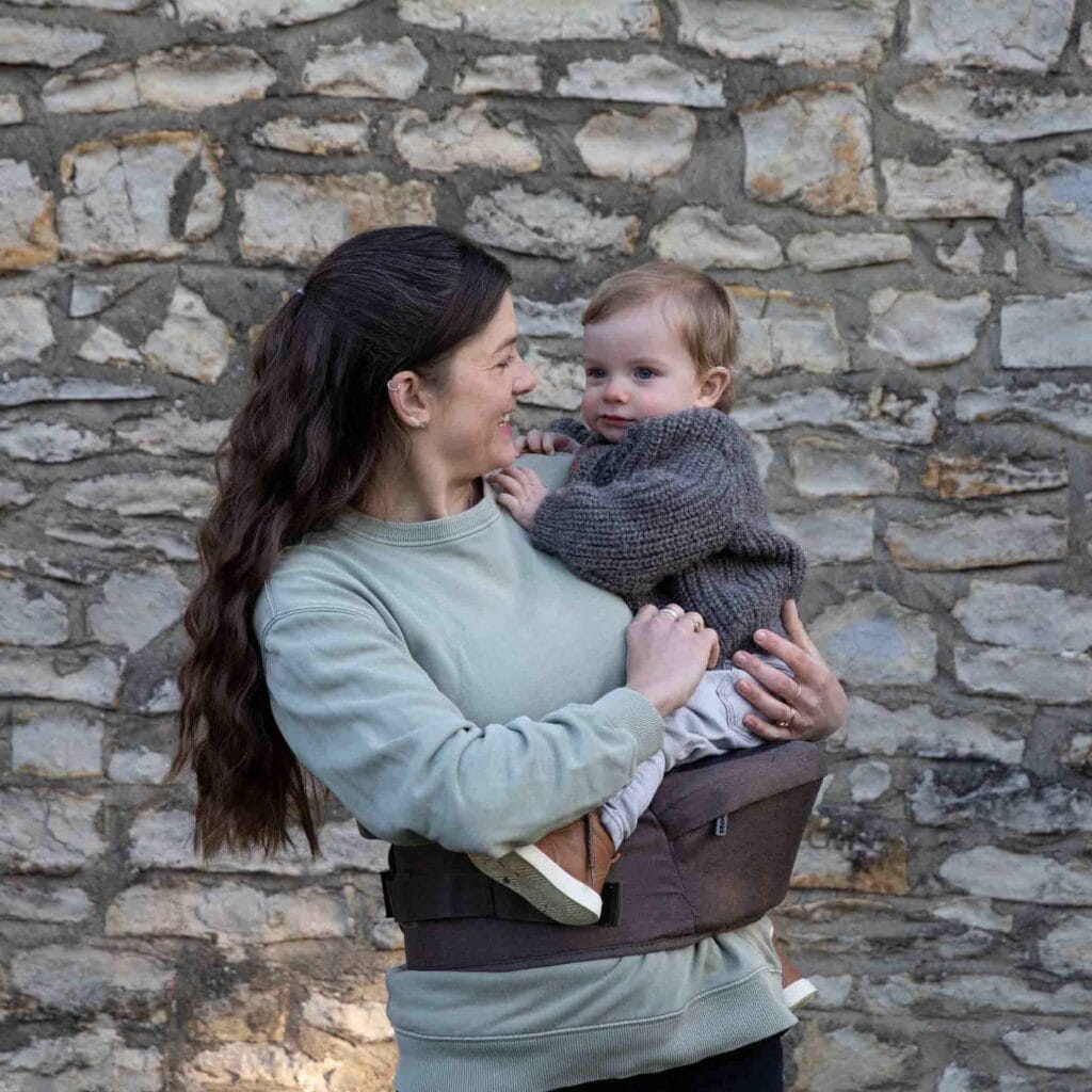 Mother holding baby on the Mocha Hipseat with brick wall in background