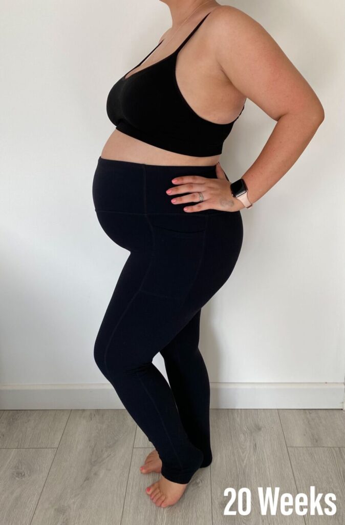 A pregnant lady stood against a white wall wearing black leggings and a black sports bra